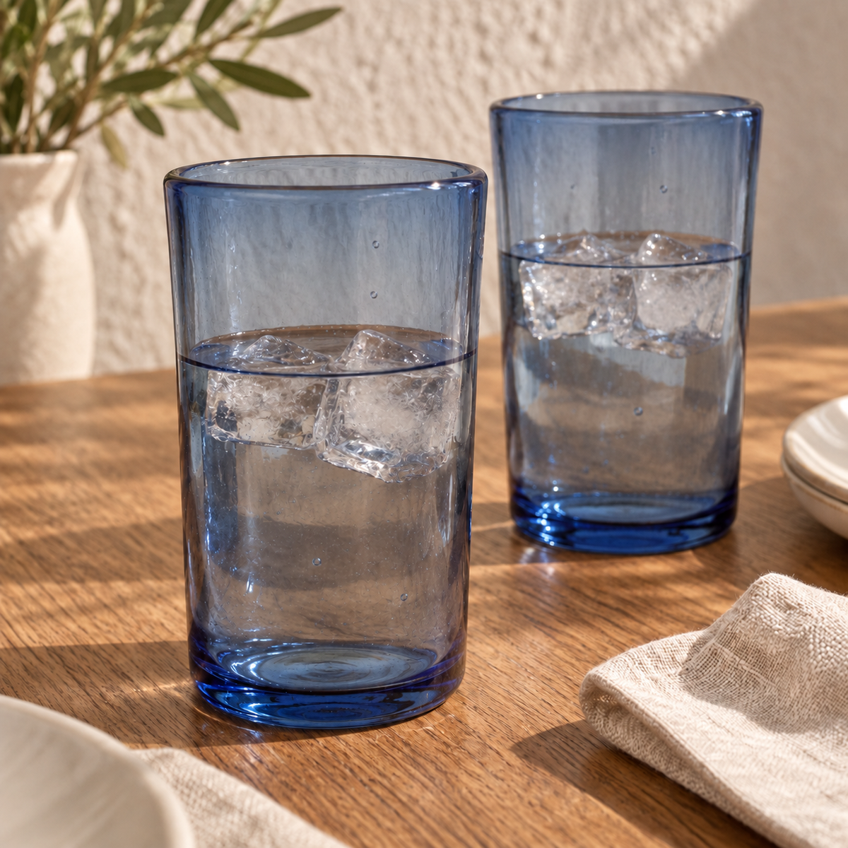 Dos vasos altos de vidrio soplado azul humo MAREY con agua y hielo en una mesa con vajilla blanca y servilleta de lino.